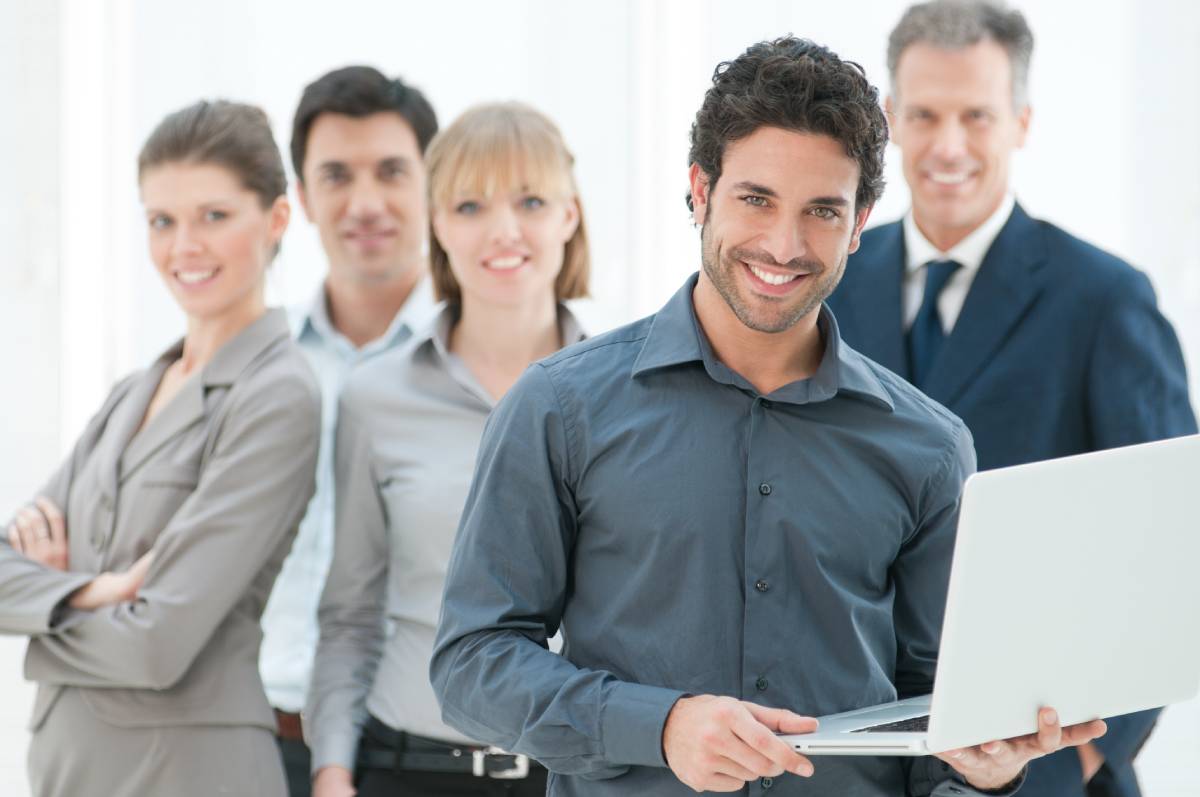 A man holding a laptop and smiling with 4 coworkers behind him smiling