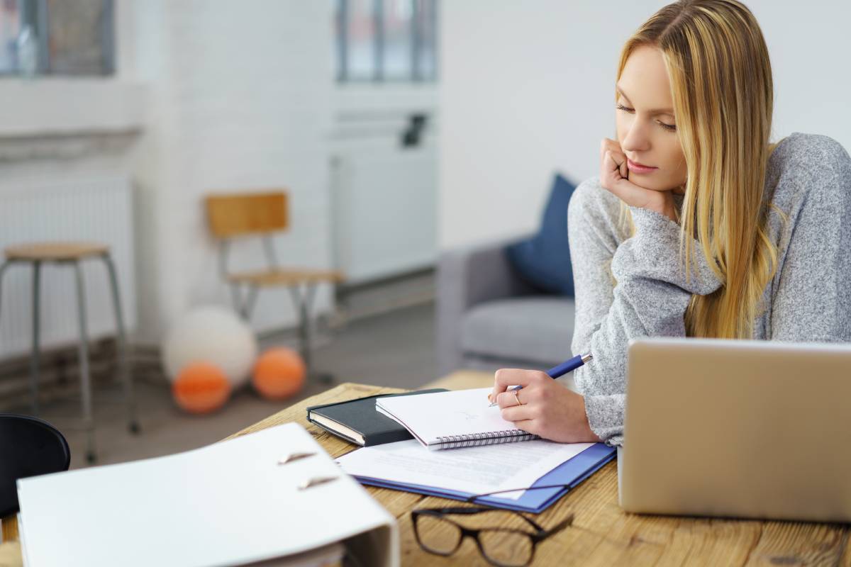 A blonde woman working from home using a laptop and writing in a notepad