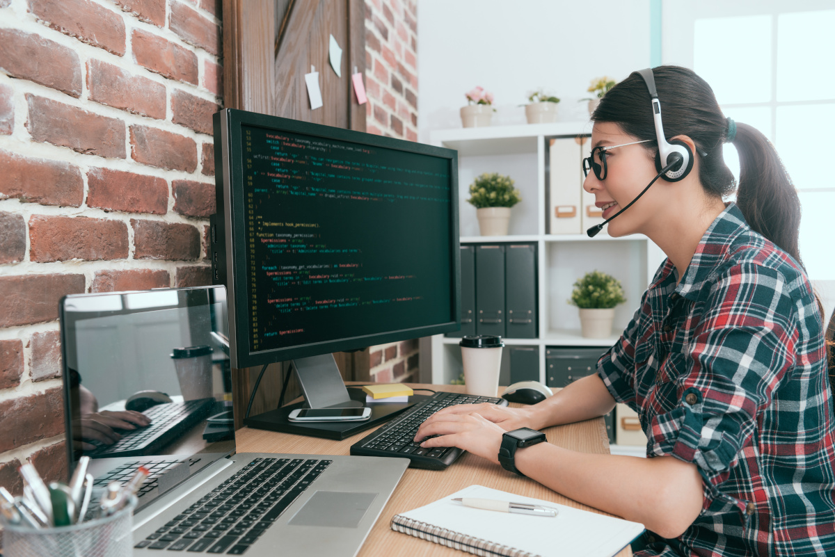 A woman wearing a headset with a mic working on code on a PC in her home office